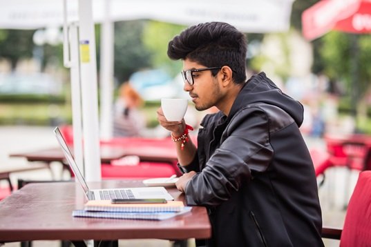 Indian man using laptop while drinking a cup coffee, outdoor street cafe